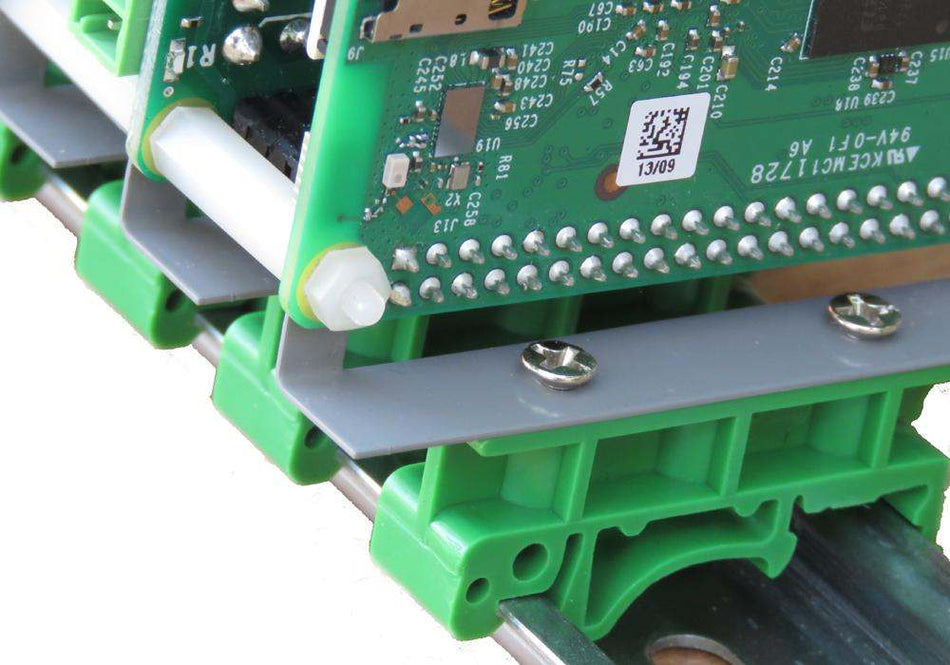Close-up of Raspberry Pi mounted perpendicularly on green plastic DIN-RAIL brackets with metal screws on a gray steel plate, showing electronic circuit board and mounting hardware.